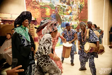 Guests in indigo and pink adire dance with drummers inside the exhibition hall.