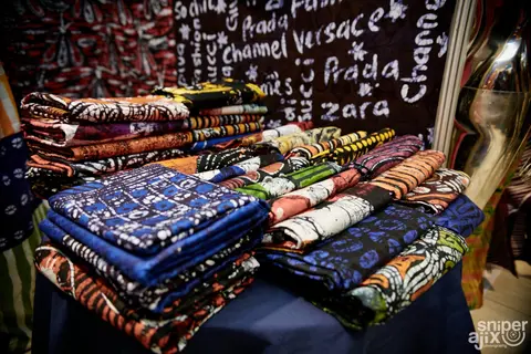 Adire textiles piled beneath hand-written designer names on a stall.
