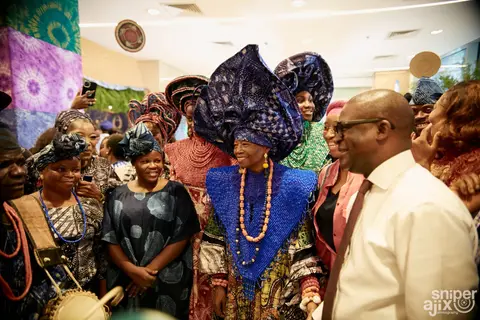 Women in elaborate indigo gele and coral beads greet guests.