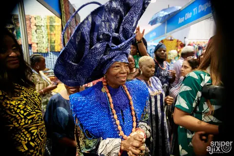A woman in an indigo gele and coral beads smiles among the crowd.