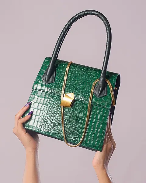 Two hands hold up an emerald croc-effect handbag with a gold clasp against lilac.