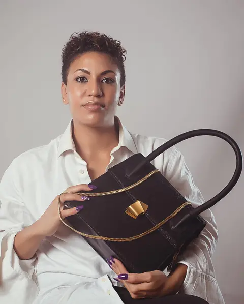 A woman with a short crop in a white shirt holds a black leather handbag.