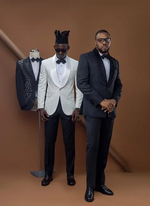 Two models in black and white tuxedos pose beside a tailoring mannequin on a warm brown set.