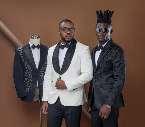 Three men in tuxedos stand beside a mannequin wearing an embellished jacket against a brown studio backdrop.