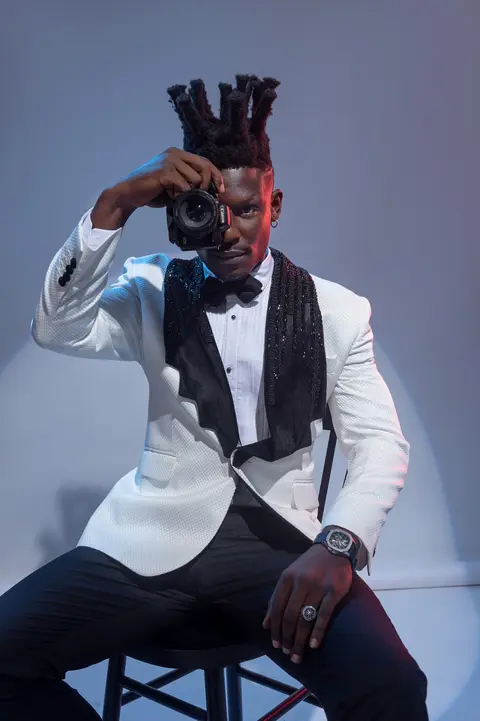 A model in a white tuxedo jacket holds a camera to his eye, seated on a wooden chair against a pale blue studio backdrop.