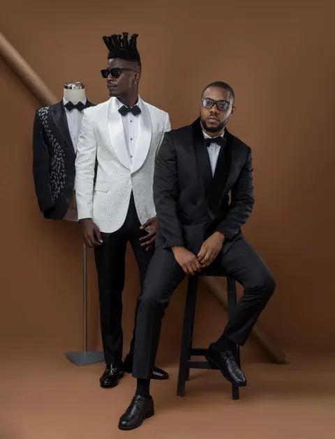 Two men in formal suits, one seated on a stool, against a brown studio wall.