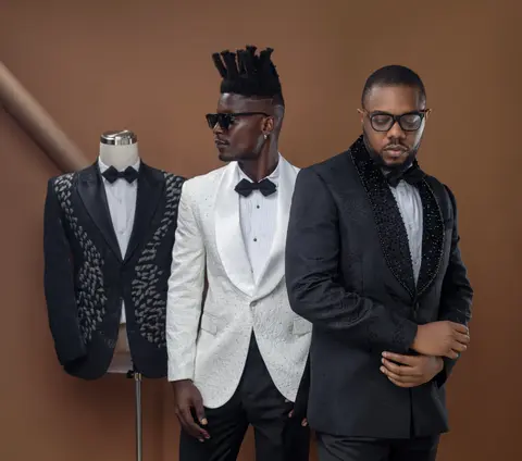 Two men in white and black tuxedo jackets pose beside an embellished mannequin.
