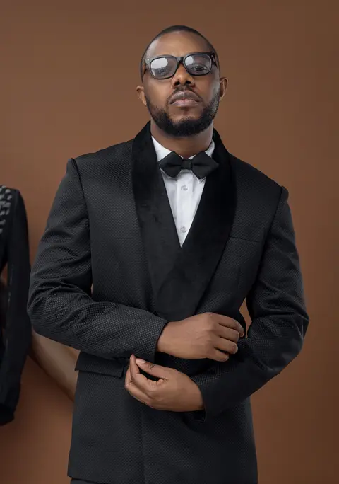 A man in glasses and a black velvet shawl-collar tuxedo adjusts his cuff.