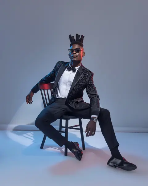 A model in a black embellished tuxedo sits on a spindle chair in cool blue light.