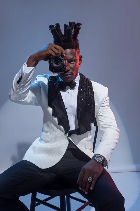 A model in a white tuxedo jacket raises a camera to his eye, seated in blue light.