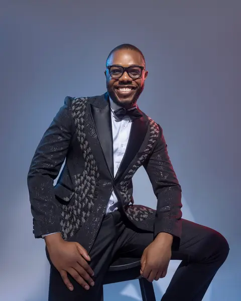 A smiling man in a black embellished tuxedo sits forward in blue studio light.