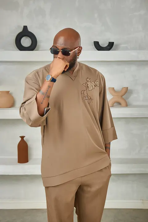 A man in a tan embroidered kaftan and sunglasses, hand at his chin, in a minimal room.