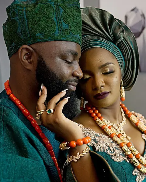 A couple in emerald aso-oke and coral beads, cheek to cheek, eyes closed.