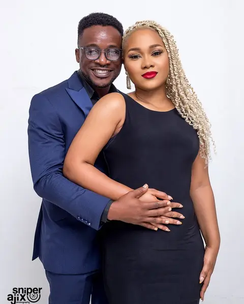 A man in a navy suit and glasses holds a woman with platinum braids in a black dress.