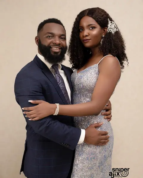 A man in a navy suit embraces a woman in a silver sequinned gown against a cream backdrop.
