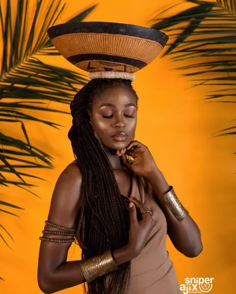 A woman with long locs balances a woven calabash on her head against orange and palm leaves.