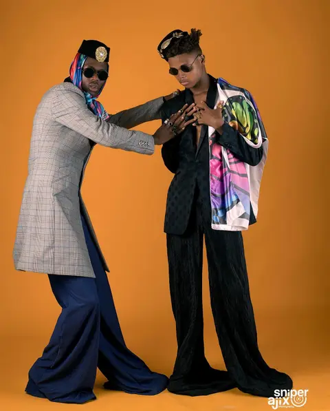 Two men in tailored suits, silk scarves and round sunglasses strike poses against orange.