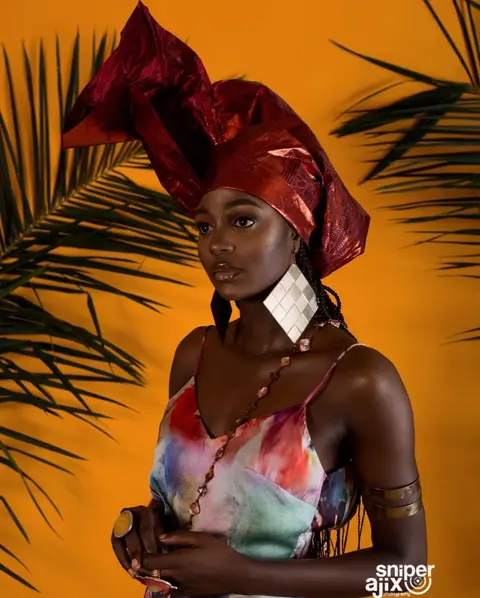 A woman in a sculpted red gele and painted silk slip against an orange backdrop with palm fronds.