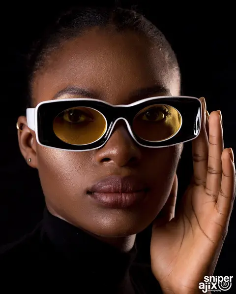 A woman in white oversized sunglasses lifts her hand to her face against black.