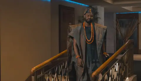 A groom in agbada and coral beads descends a staircase, from a wedding film.