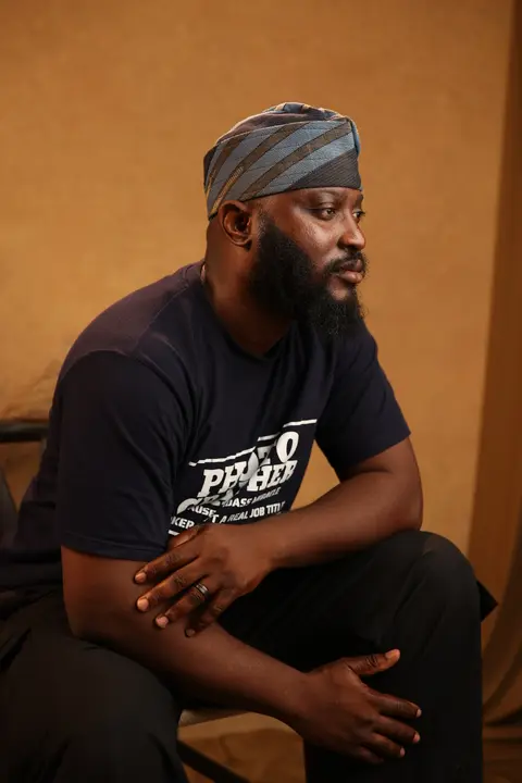 Shola Ajisegbede, founder of Sniper Ajix, in a blue and gold fila, seated against a warm amber backdrop.
