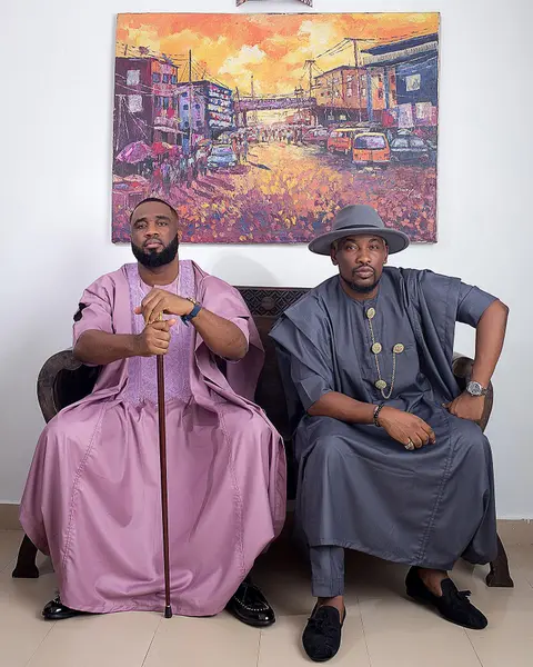Two men in lilac and grey agbada seated beneath a painting of a Lagos street.