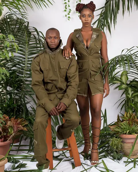 Two models in olive utility wear among potted palms, one seated on a stool.