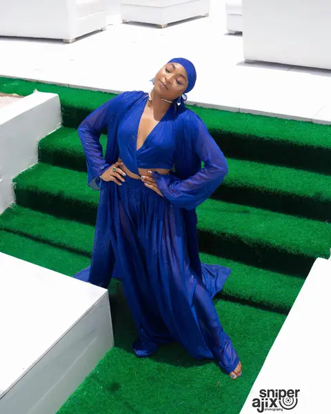 A woman in flowing cobalt chiffon and a matching head wrap poses on green-carpeted steps.