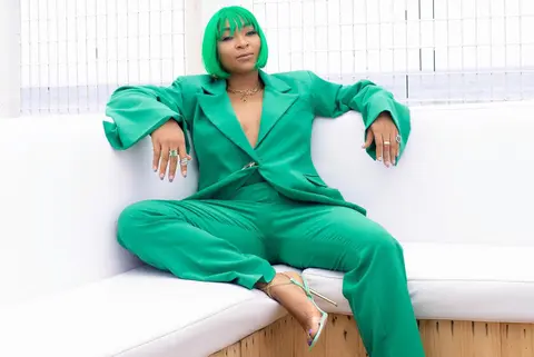 A woman with an emerald bob in a matching emerald trouser suit lounges on a white sofa.