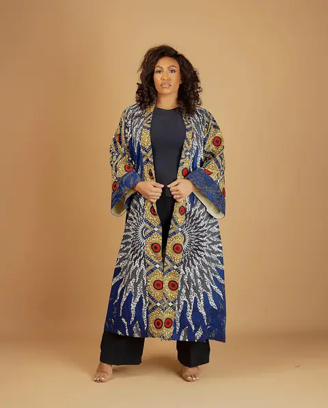 A woman in a floor-length ankara kimono coat against a tan backdrop.