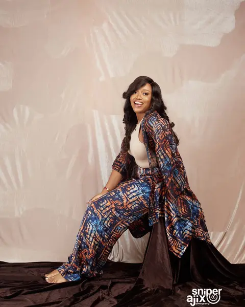 A woman in a patchwork print kimono and trousers seated on draped brown fabric.