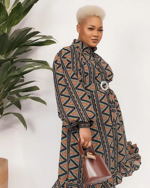 A woman with a platinum crop in a geometric print dress holds a brown leather bag beside a plant.