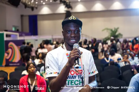 A young man speaks into a microphone in a packed hall.