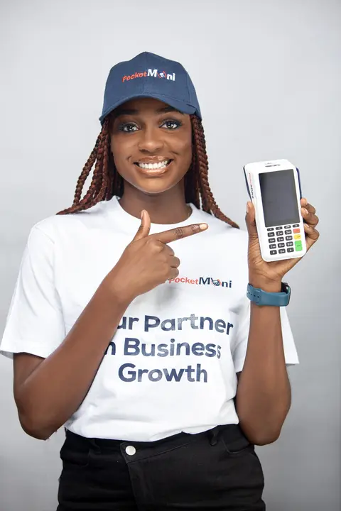 A smiling woman in a PocketMoni cap and tee points to a POS terminal.