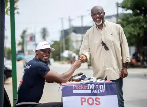 An older customer shakes hands with a PocketMoni agent at a POS stand.