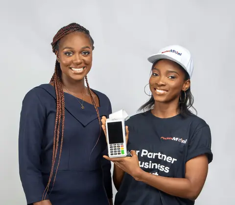 Two women smile as one holds out a PocketMoni terminal.