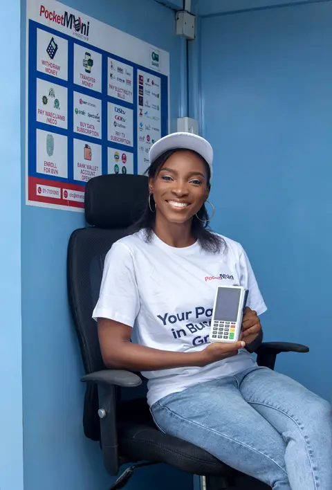 A woman sits inside a PocketMoni kiosk holding a terminal.