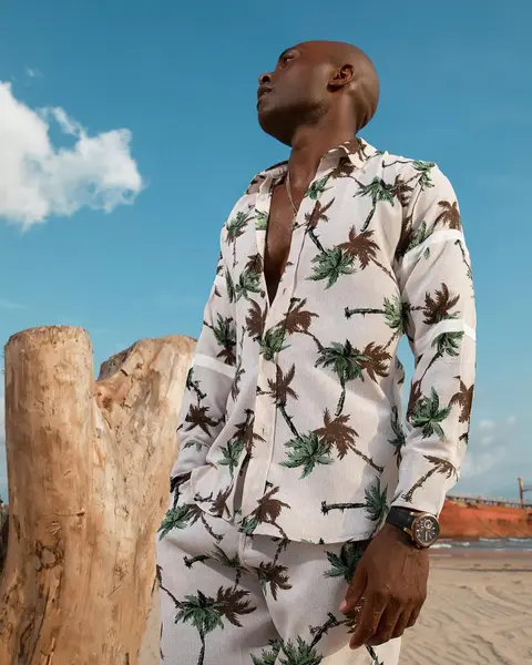A man in a palm-print shirt set looks skyward beside a driftwood trunk on the beach.