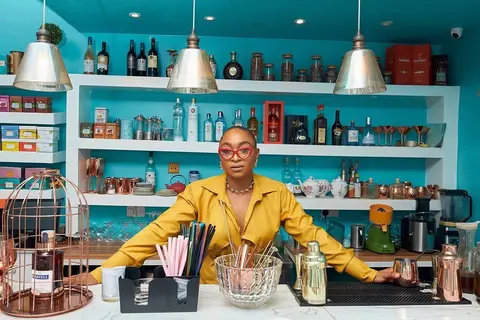 A woman in mustard leans on the counter of a teal café bar lined with bottles.
