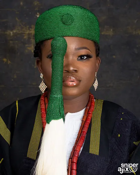 A woman in a green beaded cap with a tassel over one eye, coral beads and aso-oke.