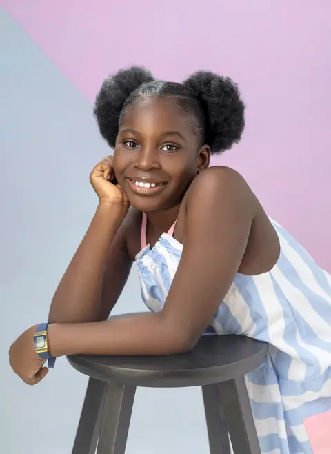 A smiling girl with two puff buns leans on a stool in a striped dress against pink and blue.