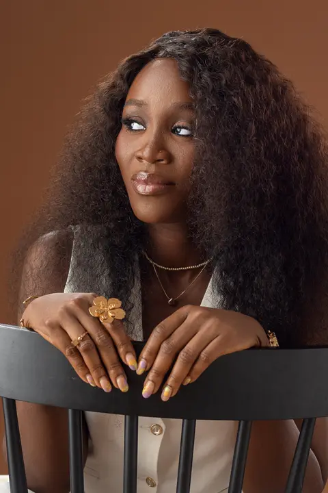 A woman with long curly hair rests her hands on a chair back against a brown backdrop.