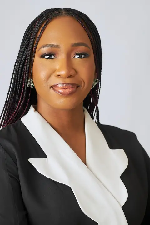 A businesswoman with long braids in a black blazer with a white lapel smiles at the camera.