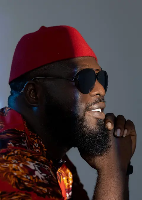 A bearded man in a red cap and dark sunglasses, chin on his hand, laughing.