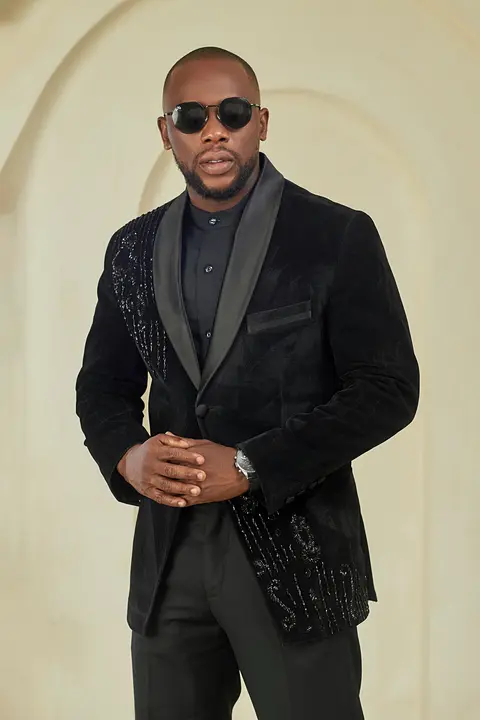 A man in a black velvet jacket and sunglasses stands before a cream arched wall.