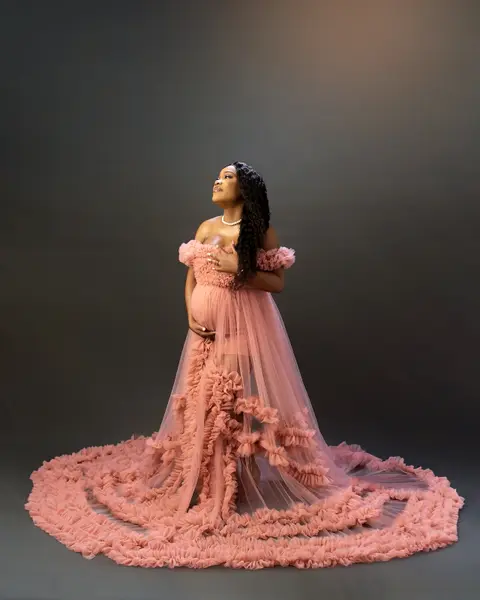 An expectant mother in a coral tulle gown with a long ruffled train, against charcoal grey.