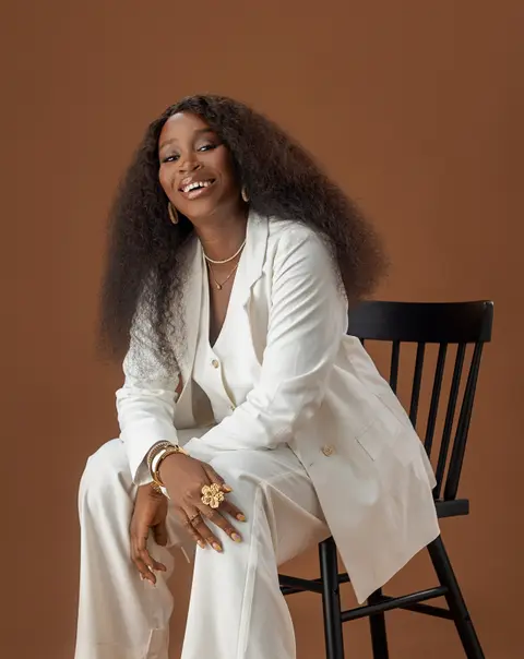 A woman in a white suit sits on a black spindle chair against a brown backdrop.