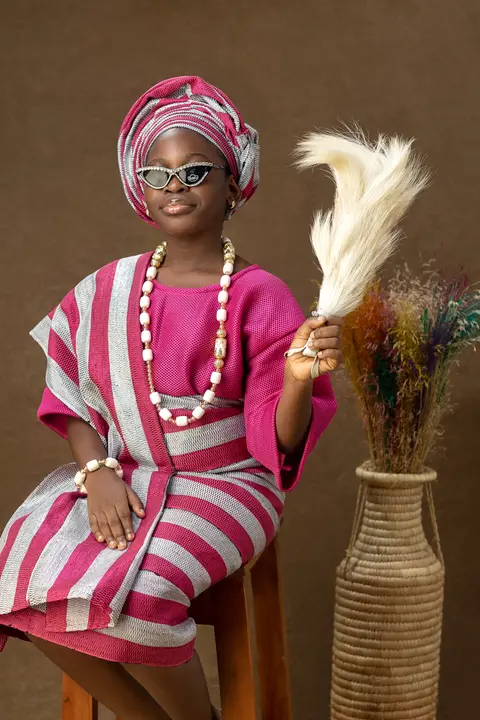 A girl in pink and white striped aso-oke, gele and coral beads holds a pampas fan beside dried flowers.