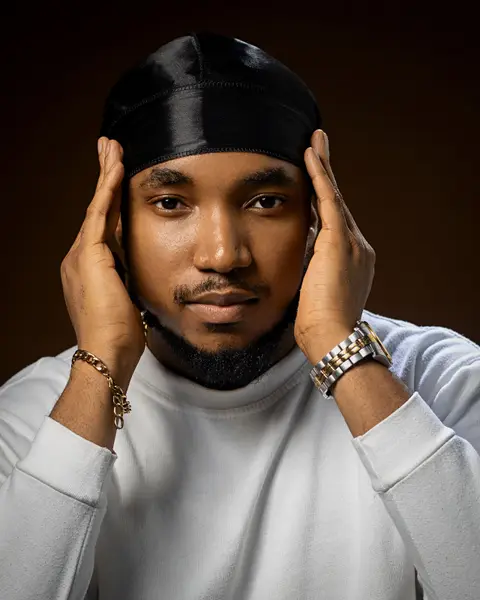 A man in a black durag and white jumper frames his face with his hands against brown.