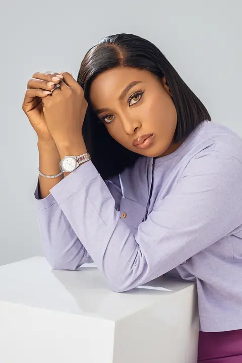 A woman in lilac leans on a white block with her hands at her chin against pale grey.
